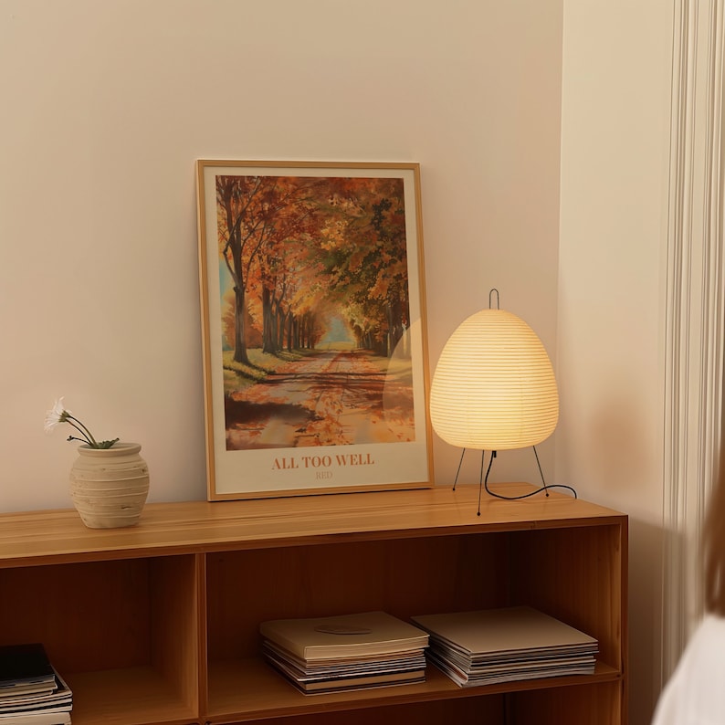 May include: A wooden shelf unit with a framed poster of an autumnal scene with the text "ALL TOO WELL (10 Minute Version)" and a white, round, hanging lamp with a black cord and stand.