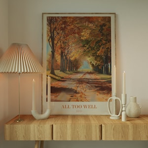 May include: A wooden table with two drawers and a white ceramic vase with a brown ceramic candle holder with two white candles. A framed poster with a painting of a path through a forest of trees with fall foliage and the text "ALL TOO WELL RED" is on the wall above the table.