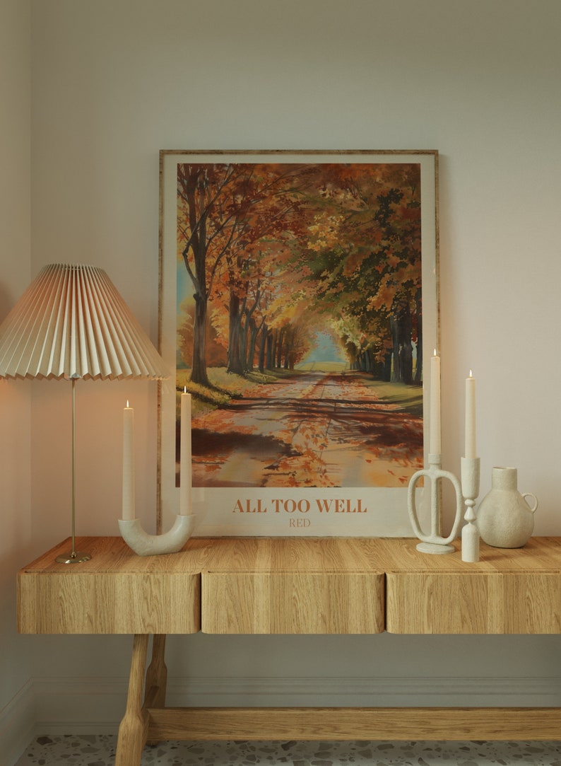 May include: A wooden table with two drawers and a white ceramic vase with a brown ceramic candle holder with two white candles. A framed poster with a painting of a path through a forest of trees with fall foliage and the text "ALL TOO WELL RED" is on the wall above the table.