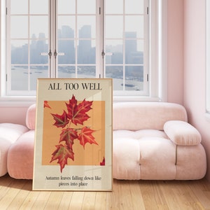 May include: A framed poster with the text "ALL TOO WELL" and a picture of red maple leaves against a light orange background. The text below the image reads "Autumn leaves falling down like pieces into place."