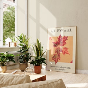 May include: A framed print with the text "ALL TOO WELL" and a botanical illustration of red maple leaves on a light yellow background. The text below the illustration reads "Autumn leaves falling down like pieces into place."