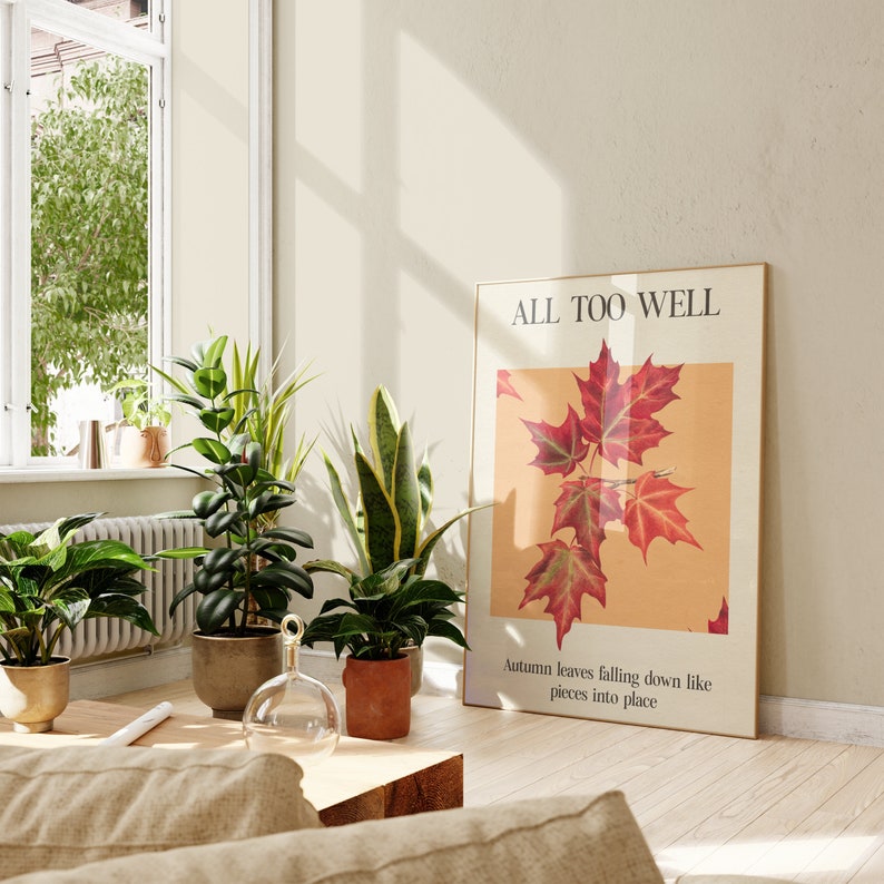 May include: A framed print with the text "ALL TOO WELL" and a botanical illustration of red maple leaves on a light yellow background. The text below the illustration reads "Autumn leaves falling down like pieces into place."
