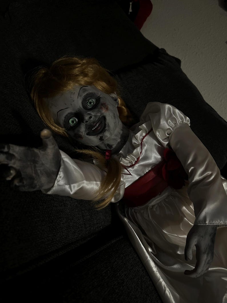 May include: A creepy doll with blonde pigtails, green eyes, and a sinister smile. The doll wears a white satin dress with red accents. The doll's skin has a pale, gray appearance, and its outstretched hand adds to the unsettling effect.