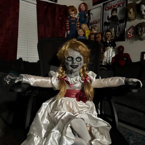 May include: A creepy Annabelle doll with blonde pigtails, wearing a white satin dress with a red sash and a red rose. The doll is seated in a black chair, with other horror movie dolls and posters in the background.