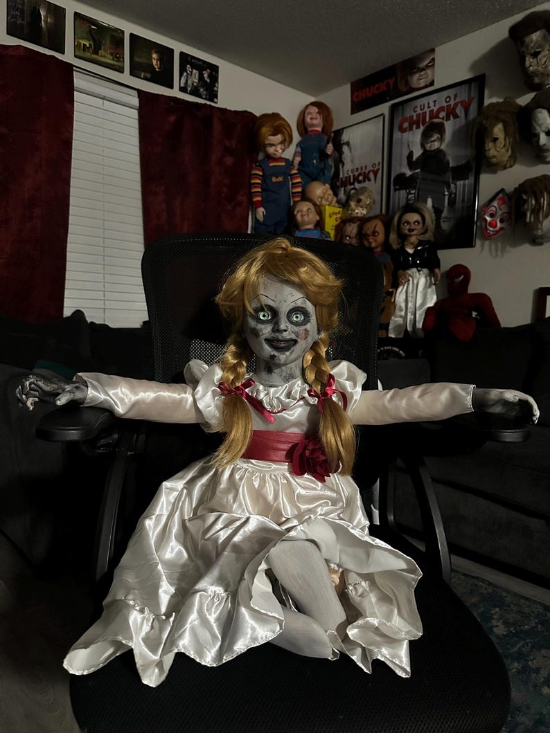 May include: A creepy Annabelle doll with blonde pigtails, wearing a white satin dress with a red sash and a red rose. The doll is seated in a black chair, with other horror movie dolls and posters in the background.