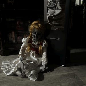 May include: A seated Annabelle doll with a pale face, blonde pigtails, and a white satin dress with a red bow. The doll is in front of a black box with the name "ANNABELLE" printed on it. The background is dark.