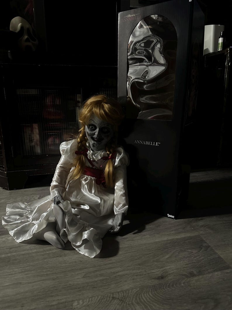 May include: A seated Annabelle doll with a pale face, blonde pigtails, and a white satin dress with a red bow. The doll is in front of a black box with the name "ANNABELLE" printed on it. The background is dark.