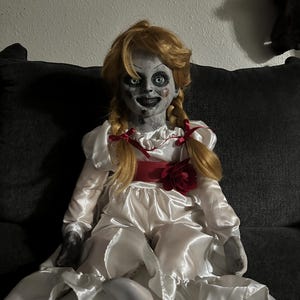 May include: A doll with a pale face, green eyes, and blonde pigtails. The doll is wearing a white satin dress with a red ribbon and a red flower. The doll is sitting on a dark gray couch.