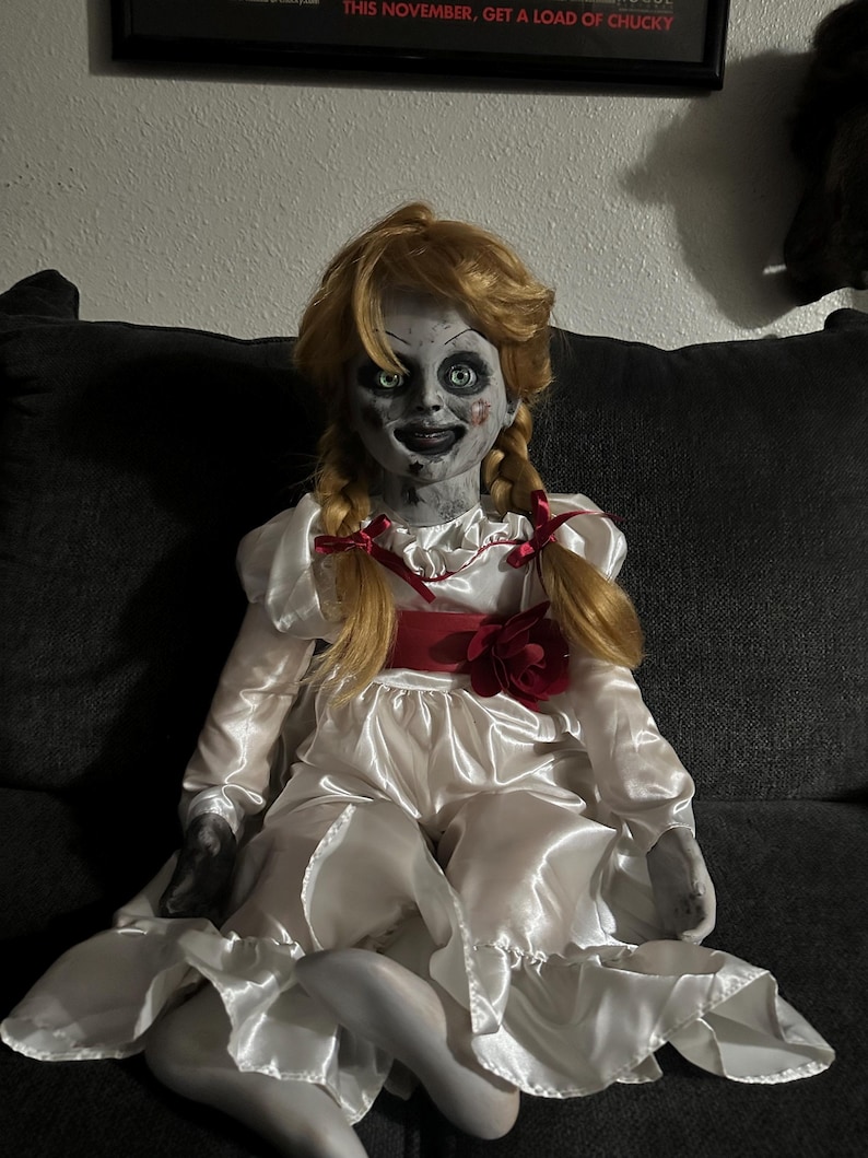 May include: A doll with a pale face, green eyes, and blonde pigtails. The doll is wearing a white satin dress with a red ribbon and a red flower. The doll is sitting on a dark gray couch.