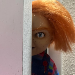Op de afbeelding: Een pop met oranje haar en blauwe ogen die achter een roze en witte muur vandaan gluurt. De pop heeft sproeten en draagt een blauwe overall met rode knopen en een gestreept shirt. De uitdrukking van de pop is ondeugend.
