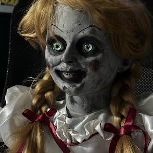 May include: A creepy doll with blonde pigtails and glowing green eyes. The doll has a pale face with dark markings and a sinister smile. It wears a white dress with a red ribbon. A horror-themed collectible.