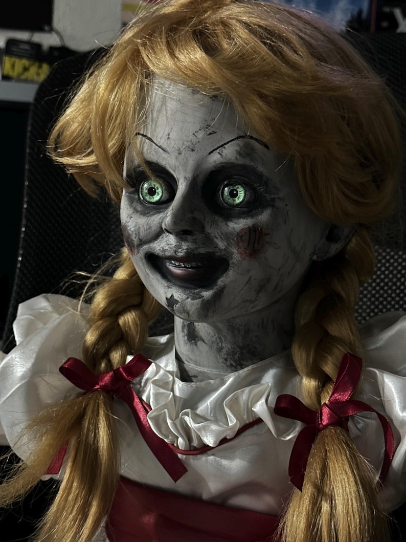 May include: A creepy doll with blonde pigtails and glowing green eyes. The doll has a pale face with dark markings and a sinister smile. It wears a white dress with a red ribbon. A horror-themed collectible.
