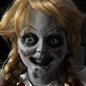 May include: A close-up of a doll with blonde hair styled in pigtails. The doll has a pale face with dark eye makeup and green eyes. Red accents are visible on the cheeks and a red bow is tied at the base of the doll's white dress.