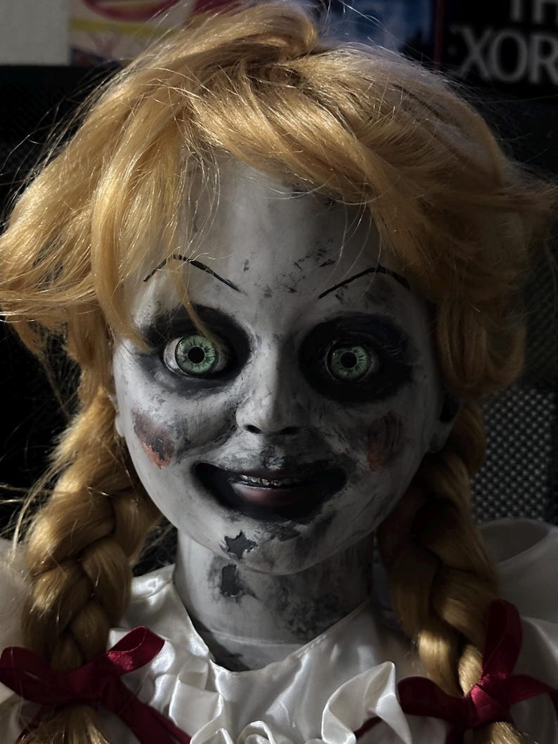 May include: A close-up of a doll with blonde hair styled in pigtails. The doll has a pale face with dark eye makeup and green eyes. Red accents are visible on the cheeks and a red bow is tied at the base of the doll's white dress.