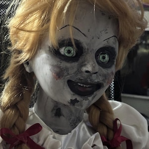 May include: A creepy doll with blonde pigtails and green eyes. The doll has a pale face with dark eye makeup and a sinister smile. It wears a white dress with red bows.