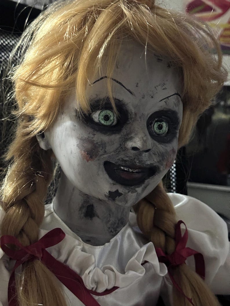 May include: A creepy doll with blonde pigtails and green eyes. The doll has a pale face with dark eye makeup and a sinister smile. It wears a white dress with red bows.