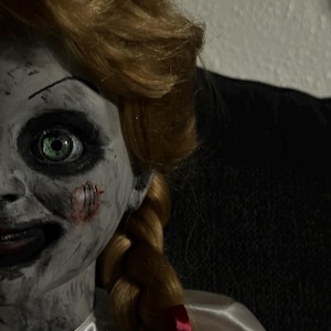 May include: A close-up of a doll with pale skin, green eyes, and blonde hair styled in a braid. The doll has a sinister expression with a stitched wound on its cheek. It wears a white satin dress with a red bow.