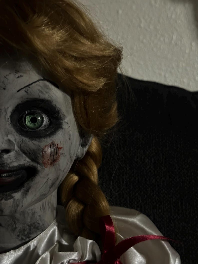 May include: A close-up of a doll with pale skin, green eyes, and blonde hair styled in a braid. The doll has a sinister expression with a stitched wound on its cheek. It wears a white satin dress with a red bow.