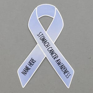 May include: A light blue ribbon with the words "Stomach Cancer Awareness" printed on it. The ribbon also has the words "Name Here" printed on it. The ribbon is on a gray background.
