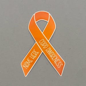 May include: An orange awareness ribbon sticker with a white border. The text "COPD AWARENESS" is printed in white on the right side of the ribbon, and the text "NAME HERE" is printed in white on the left side of the ribbon.