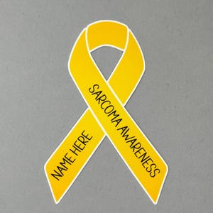 May include: A yellow awareness ribbon with the words "SARCOMA AWARENESS" and "NAME HERE" printed on it. The ribbon is on a gray background. The ribbon is a symbol of support for sarcoma awareness.