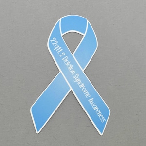 May include: A light blue ribbon with the text "22q11.2 Deletion Syndrome Awareness" printed on it.