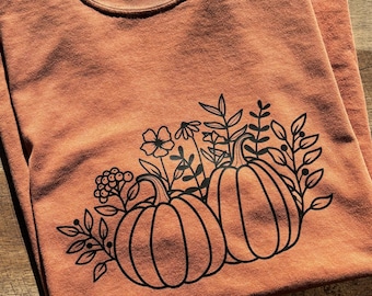 May include: A rust-colored t-shirt with a black graphic of two pumpkins with stems and leaves. The pumpkins are surrounded by black floral designs.