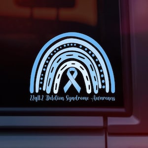 May include: A blue and white rainbow with a blue ribbon in the center. The text "22q11.2 Deletion Syndrome - Awareness" is below the rainbow.