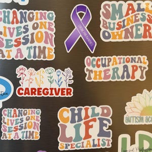 May include: A collection of colorful stickers with various text and designs. The stickers include phrases like "Changing Lives One Session at a Time," "Small Business Owner," and "Child Life Specialist." There is also a purple ribbon sticker with the text "Chronic Pain Awareness."