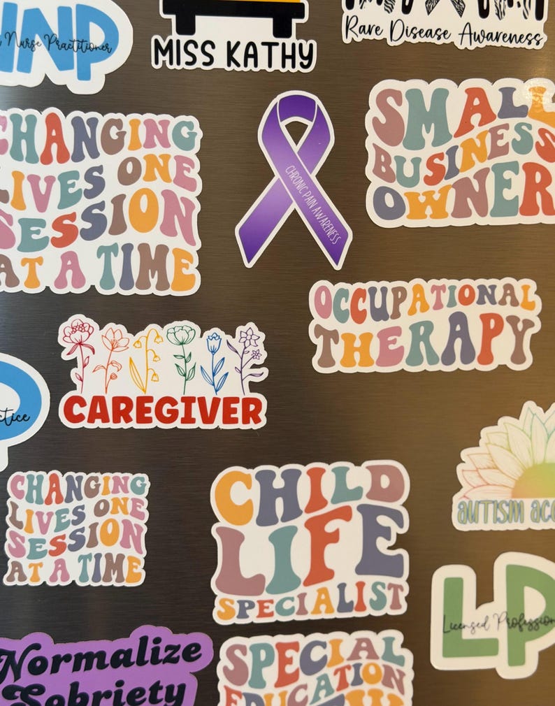 May include: A collection of colorful stickers with various text and designs. The stickers include phrases like "Changing Lives One Session at a Time," "Small Business Owner," and "Child Life Specialist." There is also a purple ribbon sticker with the text "Chronic Pain Awareness."