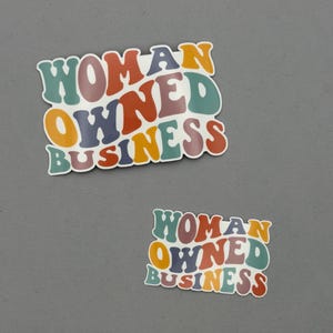 May include: Two stickers with the text "WOMAN OWNED BUSINESS" in a retro font. The text is in various colors, including orange, green, purple, and red. The stickers are on a gray background.