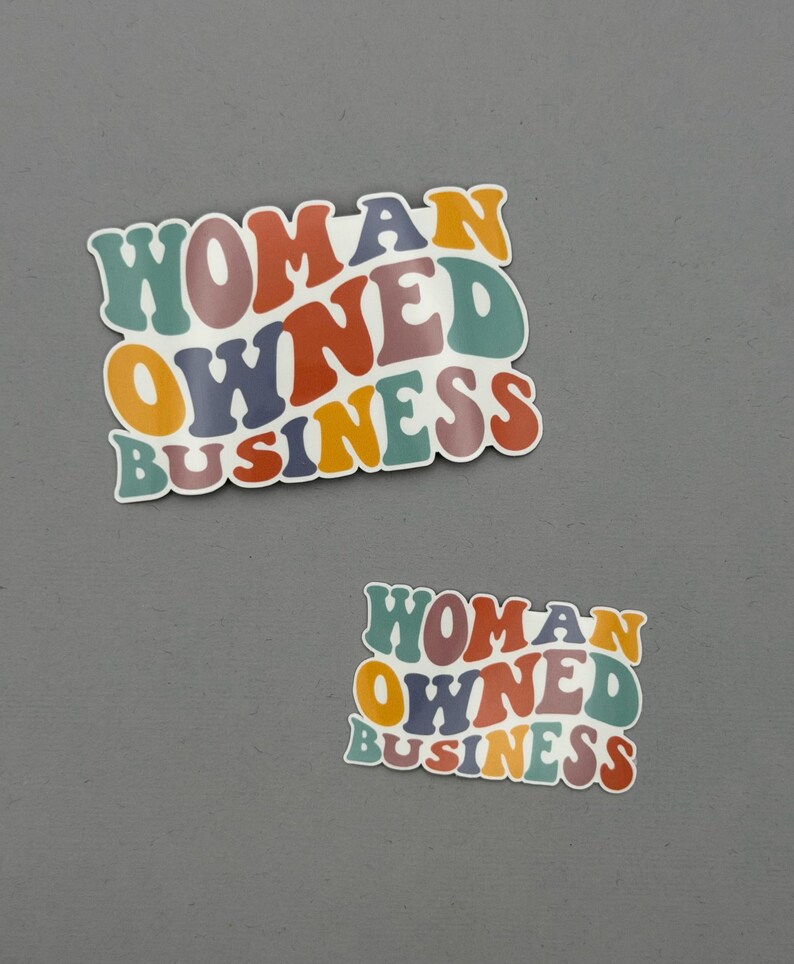 May include: Two stickers with the text "WOMAN OWNED BUSINESS" in a retro font. The text is in various colors, including orange, green, purple, and red. The stickers are on a gray background.