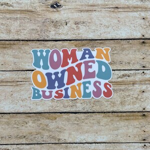 May include: A colorful sticker with the words "WOMAN OWNED BUSINESS" in a retro font. The text is in shades of blue, orange, purple, and red, with a white outline. The sticker is on a wooden background.