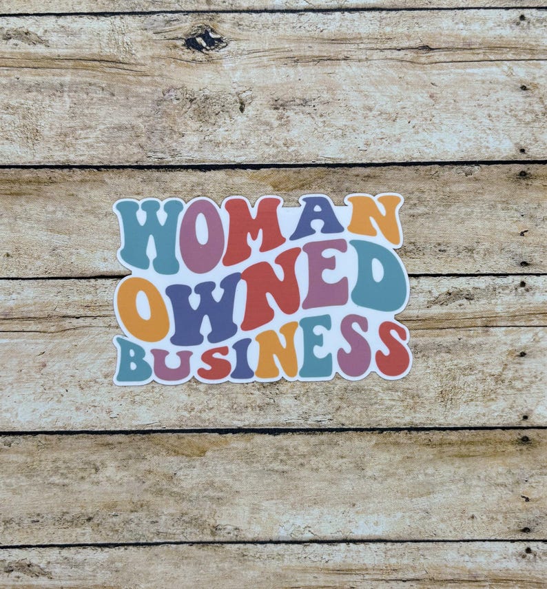 May include: A colorful sticker with the words "WOMAN OWNED BUSINESS" in a retro font. The text is in shades of blue, orange, purple, and red, with a white outline. The sticker is on a wooden background.