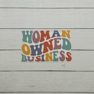 May include: A sticker with the words "WOMAN OWNED BUSINESS" in a retro font. The text is in various colors, including orange, teal, purple, and yellow. The sticker is on a white wooden background.