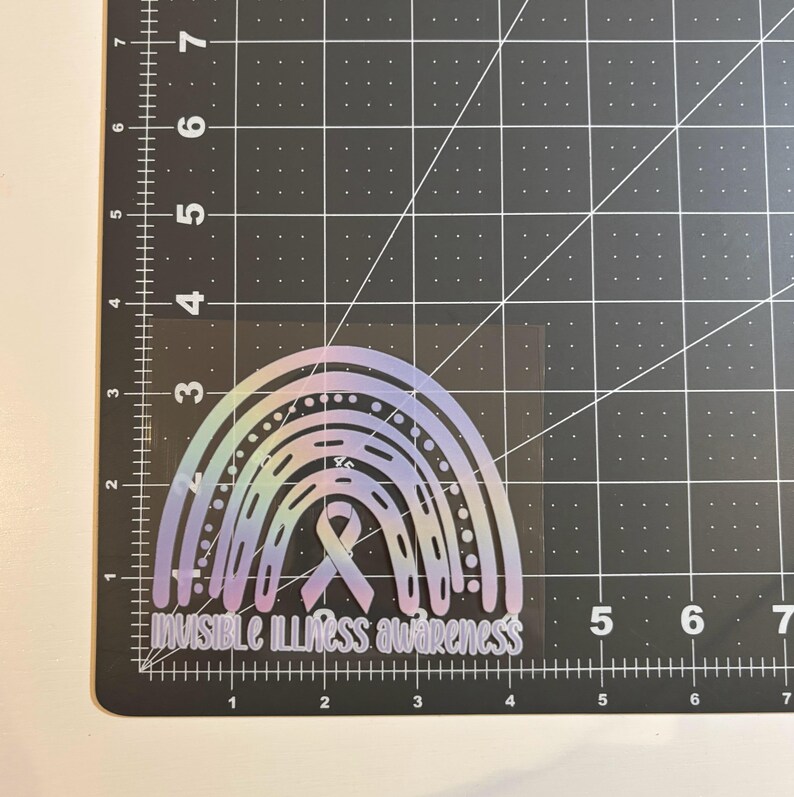 May include: A holographic rainbow sticker with the text "Invisible Illness Awareness" in a script font. The sticker features a ribbon in the center of the rainbow design. The sticker is on a black grid surface.