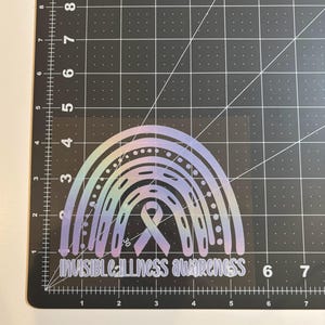 May include: A holographic rainbow sticker with the text "INVISIBLE ILLNESS AWARENESS". The sticker features a rainbow design with a ribbon in the center. The sticker is on a black cutting mat.