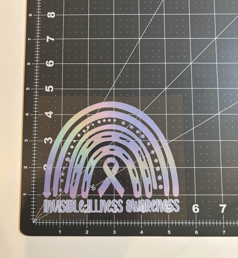 May include: A holographic rainbow sticker with the text "INVISIBLE ILLNESS AWARENESS". The sticker features a rainbow design with a ribbon in the center. The sticker is on a black cutting mat.