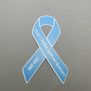 May include: Light blue ribbon with the text "22q11.2 Deletion Syndrome Awareness" and "NAME HERE" printed on it.