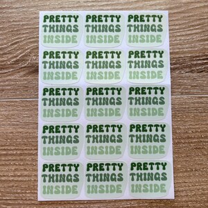 May include: A sheet of stickers with the text "PRETTY THINGS INSIDE" in a retro font. The text is in a dark green color, with a light green background. The stickers are rectangular with rounded corners.