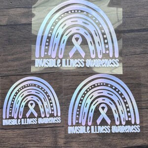 May include: Three holographic rainbow stickers with the text "Invisible Illness Awareness". Each sticker features a rainbow design with a ribbon in the center. The stickers are in different sizes and are placed on a wooden surface.