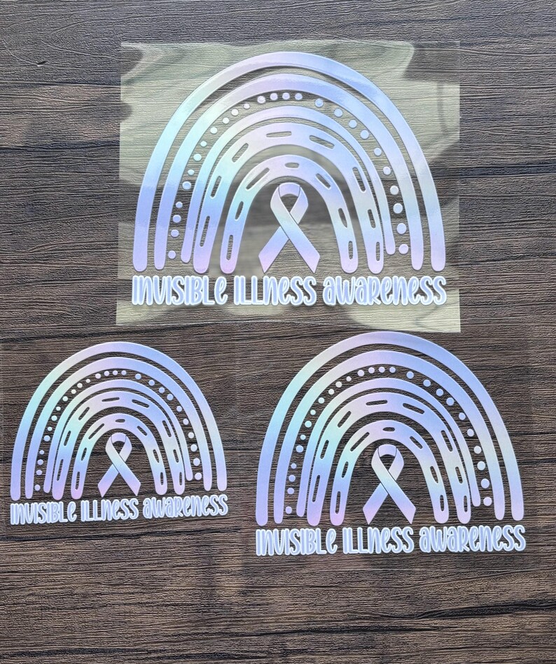 May include: Three holographic rainbow stickers with the text "Invisible Illness Awareness". Each sticker features a rainbow design with a ribbon in the center. The stickers are in different sizes and are placed on a wooden surface.