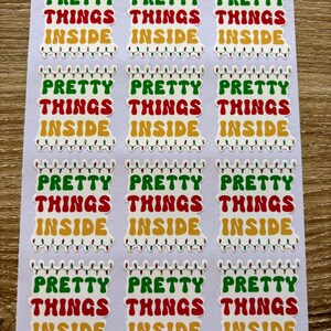 May include: Twelve white stickers with the phrase "Pretty Things Inside" in red, green, and yellow letters. The letters are outlined in white and the phrase is surrounded by a string of Christmas lights.