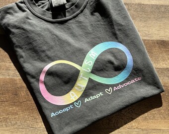 May include: A dark gray t-shirt with a rainbow infinity symbol that says "Autism" inside the symbol. Below the symbol, it says "Accept ♥ Adapt ♥ Advocate".