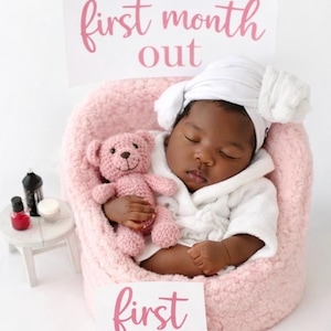 May include: A sleeping baby wearing a white robe and hat, nestled in a pink fuzzy chair, holding a pink teddy bear. The words "first month out" and "first month" are displayed in pink script on signs.