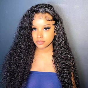 May include: A woman with long, dark curly hair wearing a blue top. The hair is styled in a long, flowing wig with a lace front.