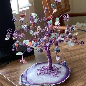 May include: A handcrafted wire tree sculpture with a purple trunk and branches, adorned with colorful gemstone leaves. The tree sits on a polished, purple-edged agate base, creating a decorative and artistic piece.