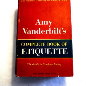 May include: A hardcover book titled "Amy Vanderbilt's Complete Book of Etiquette." The cover is primarily red with white text, a blue and gold band, and the text "The Guide to Gracious Living." The book is a guide to manners.