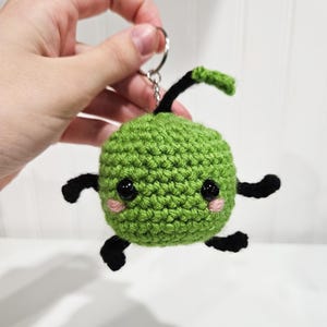 May include: Green crocheted keyring with black legs and a tail. The keyring has a cute face with black eyes and pink cheeks.