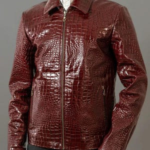 Könnte beinhalten: Eine burgunderfarbene Lederjacke mit einem Krokodilprägemuster. Die Jacke hat einen Reißverschluss und einen Stehkragen.